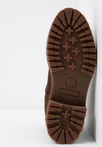 Semelle d'une botte Timberland marron avec des crampons profonds, des motifs en forme de croix et le logo de la marque visible sur un fond blanc.