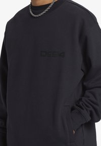 Sudadera de color azul marino oscuro, hecha de una mezcla de algodón. Presenta cuello redondo, mangas largas, un logo frontal en negro y bolsillos laterales. Textura suave.