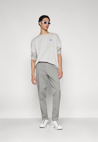 Lichtgrijze langärmige shirt met blauw logo, gecombineerd met grijze broek en witte sneakers. Accessoires omvatten witte zonnebril en een zwarte beanie.