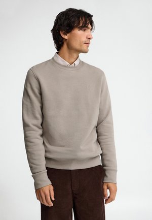 Hombre con sudadera beige claro sobre camisa blanca con cuello y pantalones marrón oscuro, de pie sobre fondo blanco, mirando hacia la derecha.
