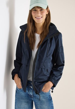 Mujer joven sonriente, que lleva una gorra azul claro, chaqueta azul marino con cremallera, camisa blanca y vaqueros azules, de pie con las manos en los bolsillos frente a una pared neutra.