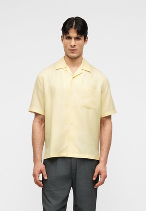 Άνδρας που στέκεται φορώντας μια ανοιχτόκίτρινη κοντομάνικη πουκάμισο με τσέπη στο στήθος και σκούρο γκρι παντελόνι, σε απλό φόντο.