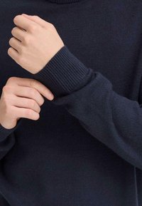 Pulover albastru închis cu mansete împletite, arătând un prim-plan cu mâini ajustând mâneca. Textură moale, croială confortabilă și design simplu.