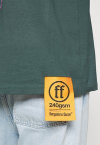 Vihreä t-paita, jossa on tekstuuripinta, oranssi etiketti mustalla tekstillä, jossa lukee "ff" ja "240gsm" helman kohdalla.