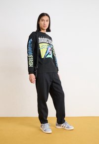 Black long-sleeve shirt with colorful graphics, paired with black pants and blue sneakers with a patterned design, set against a yellow background.