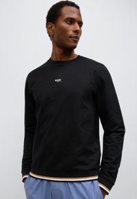 Juodas džemperis iš minkštos medžiagos, krūtinėje mažas baltas prekinis ženklas, rankogaliai su briaunelėmis ir sluoksniuotas apačia su rudos ir baltos spalvų juostelėmis.