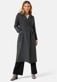 Grijze dubbelzijdige wollen jas met afgeronde revers, zijzakken en een lange, rechte pasvorm. Geddragen over zwarte broek en grijze enkellaarsjes.