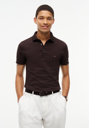 Jeune homme portant un polo noir et un pantalon blanc, mains dans les poches, souriant sur un fond blanc uni.