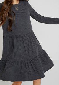 Grå ribbad klänning med långa ärmar, rund halsringning och ett lager-på-lager, utsvängd kjol. Mjuk textur med vertikala ränder och en avslappnad design.