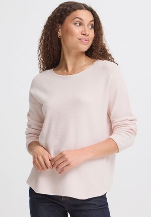 Femme aux cheveux bouclés portant un pull rose clair à manches longues et un jean foncé, regardant vers sa droite avec les mains légèrement jointes.