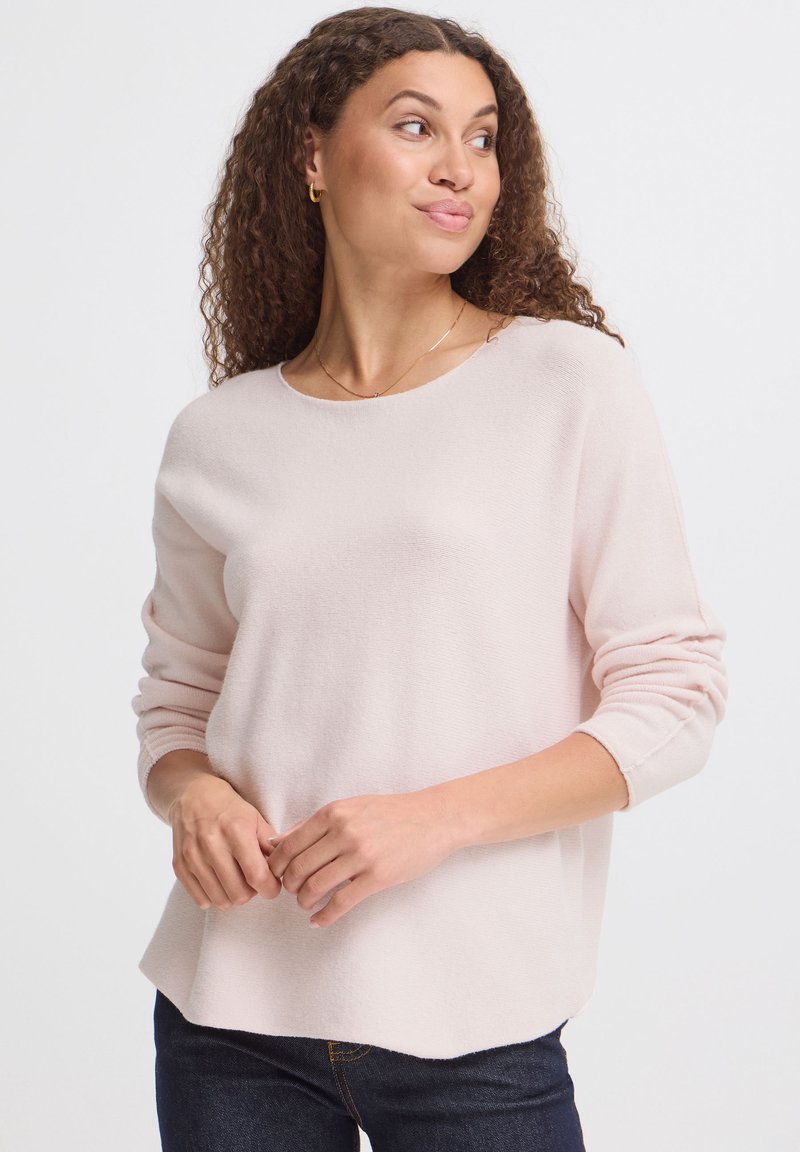 Femme aux cheveux bouclés portant un pull rose clair à manches longues et un jean foncé, regardant vers sa droite avec les mains légèrement jointes.