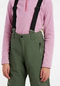 Zelene sniježne hlače s crnim podesivim naramenicama, s utorom za zatvarač na nozi, nosi se preko svijetlopinkaste polar fleece majice sa zatvaračem.