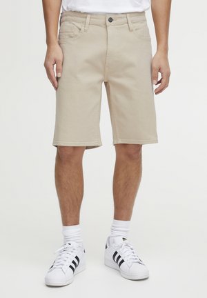 Shorts beige jusqu'aux genoux en coton, avec un design classique à cinq poches, des passants de ceinture et un système de fermeture à bouton. Portés avec des baskets blanches.