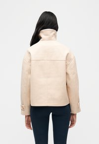 Γυναίκα με μακριά σκούρα μαλλιά γυρισμένη από πίσω φοράει μπεζ μάλλινο μπουφάν με ψηλό γιακά και μαύρο εφαρμοστό παντελόνι μπροστά σε απλό φόντο.