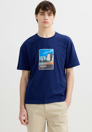 Joven con cabello oscuro que lleva una camiseta azul marino con un gráfico colorido de la playa y los acantilados de Étretat en Normandía.