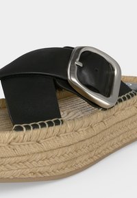 Bride de sandale en cuir noir avec grande boucle argentée sur semelle plateforme beige tissée avec couture détaillée en corde.