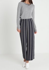 Pull gris à manches longues avec un col rond, associé à un pantalon large rayé bleu marine et blanc. Le mannequin porte des chaussures blanches à talons.