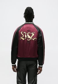 Mees, kellel on dreadsid, kandes burgundia ja musta satiinist bomberjopet, mille seljal on tikitud stiliseeritud tekst, koos mustade pükstega.