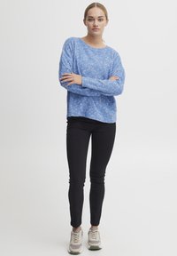 Kék, texturált pulóver hosszú ujjakkal és kerek nyakkivágással, fekete, skinny farmernadrággal és szürke sportcipővel, zöld részletekkel párosítva.