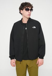 Μαύρο μπουφάν bomber με φερμουάρ μπροστά, ελαστικά μανίκια και δύο λογότυπα στο στήθος. Συνδυάζεται με μαύρη μπλούζα και πράσινο παντελόνι.