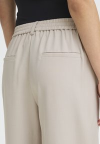 Pantalon beige avec une ceinture élastique froncée, comprenant deux poches arrière et un tissu léger et lisse.