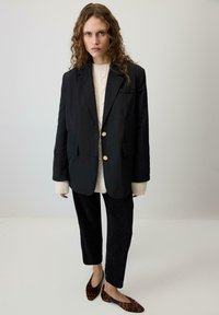 Melna pielāgota žakete ar zelta pogām, krēmkrāsas kabelēta džemperis, melni korduroja bikses un brūni leoparda raksta dzīvokļi.