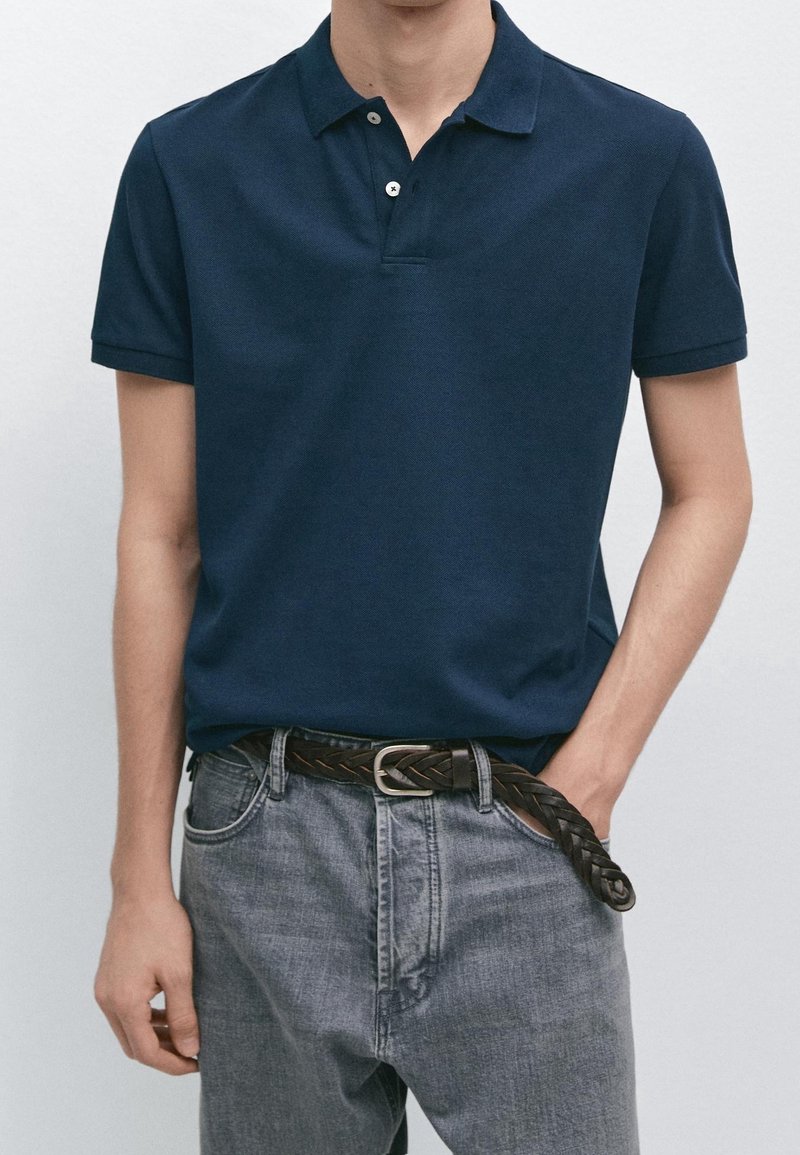 Homme portant un polo bleu foncé à manches courtes rentré dans un jean gris clair avec une ceinture tressée marron foncé, main dans la poche.
