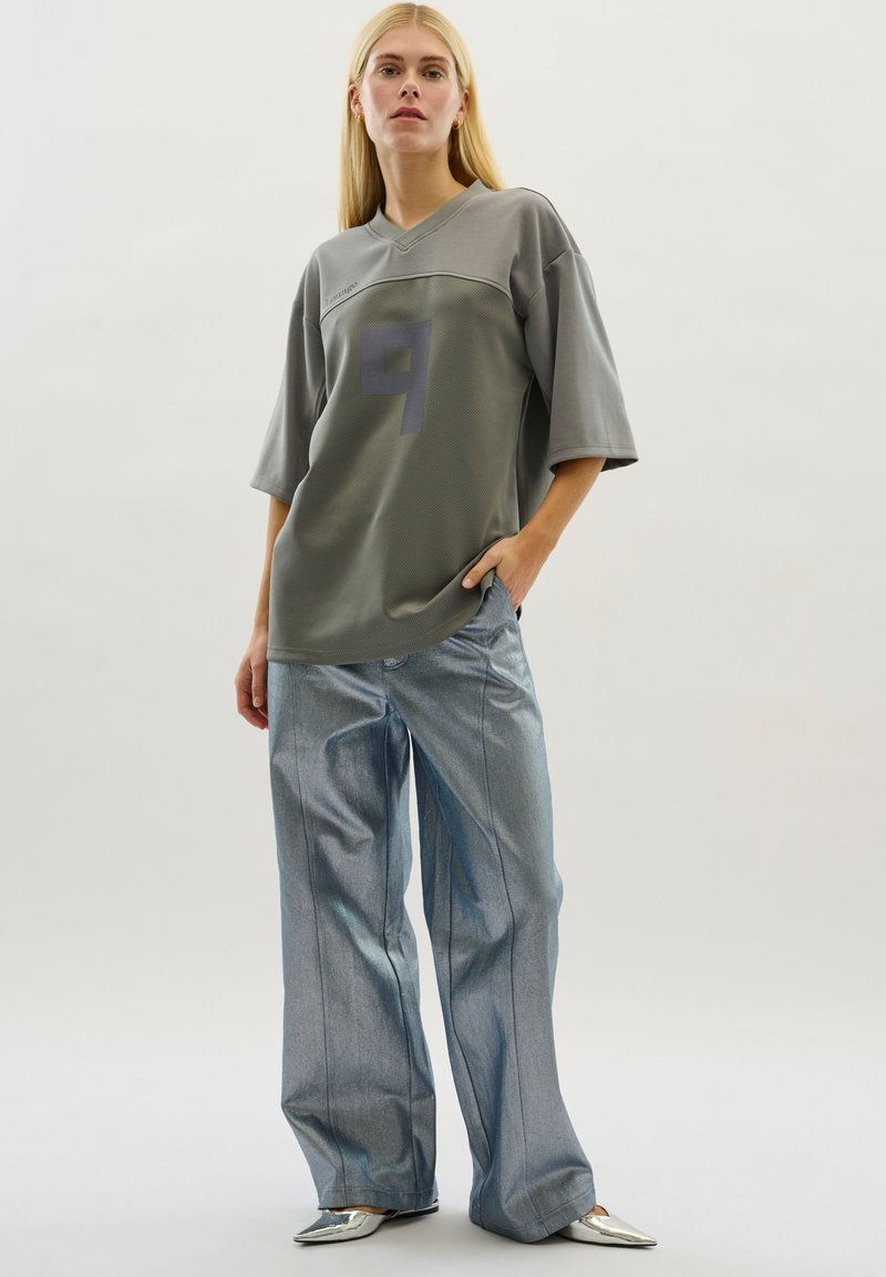 Oversized grijze trui met een v-hals en grote "9" graphic, gecombineerd met wijde metallic blauwe broeken en zilveren puntige schoenen.