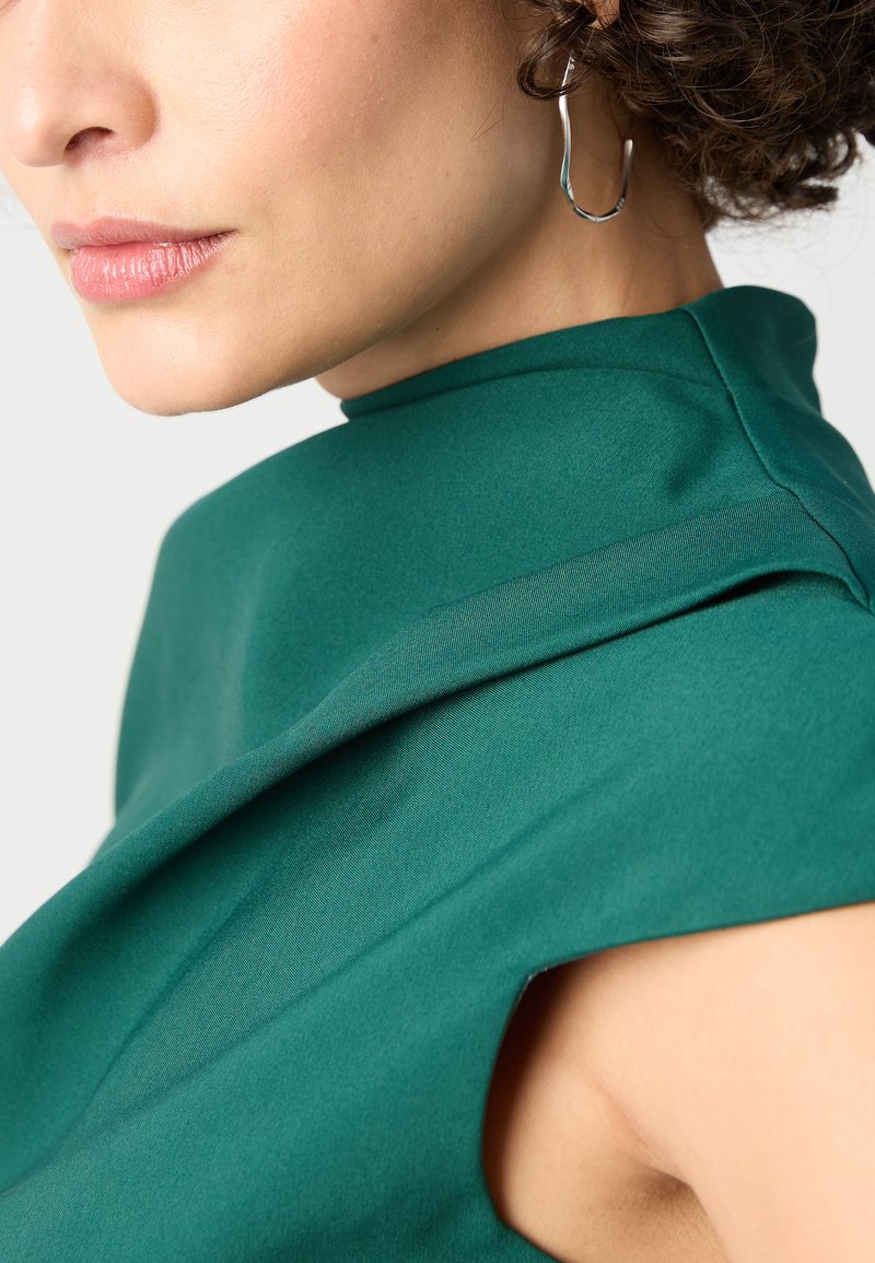 Zielona sukienka z wysokim kołnierzem, wykonana z usztywnionego materiału, z marszczonym detalem na ramieniu i wycięciami po bokach. Widoczne metalowe kolczyki w kształcie kół.