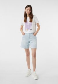 Biała koszulka z lawendową grafiką i napisem, w połączeniu z jasnoniebieskimi szortami dżinsowymi. Model nosi białe sneakersy, ręce w kieszeniach.