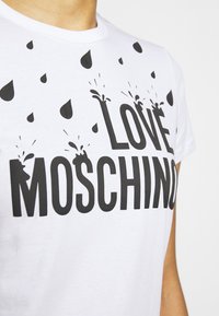 Bílá tričko s černým textem "Love Moschino" a černými grafikami kapek vody nad a kolem textu.