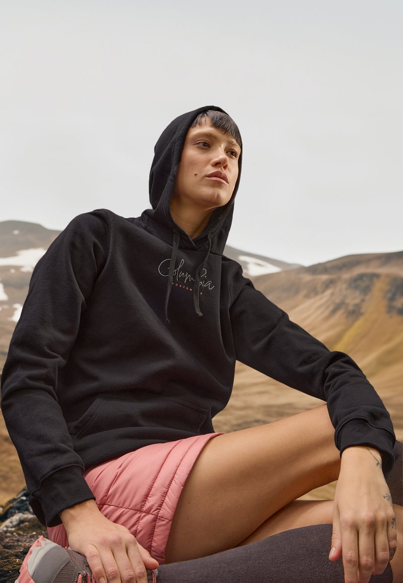 Person im Freien in schwarzem Hoodie und pinken Shorts, der auf dem steinigen Boden sitzt, mit Bergen und schneebedeckten Stellen im Hintergrund.