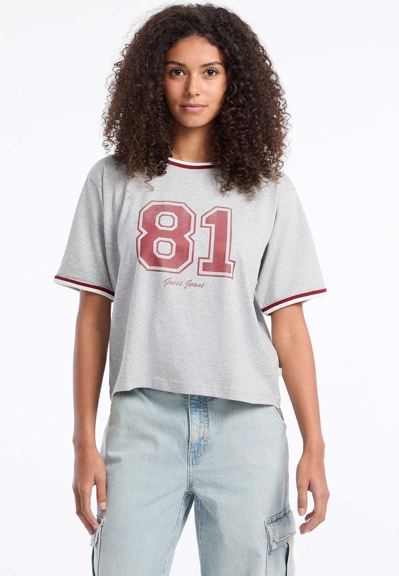 Vaalea harmaa lyhythihainen t-paita, jossa on punaiset yksityiskohdat. Paidassa on rohkea valkoinen ja viininpunainen "81" -grafiikka ja sen alla "Guess Jeans" -teksti.