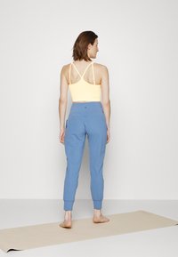 Top desportivo amarelo com alças em cruz e calças de jogging azul claro com punhos nos tornozelos, em pé sobre um tapete de ioga bege.
