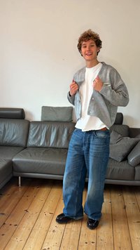 Ung person med lockigt hår som ler, klädd i en grå kofta över en vit skjorta och vida blå jeans, står på ett trägolv bredvid en grå soffa.