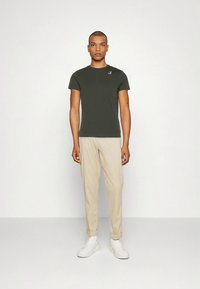 Temno zelena majica s kratkimi rokavi in rebrasto strukturo ter majhnim logotipom na prsih, kombinirana s bež zoženimi hlačami in belimi supergami.