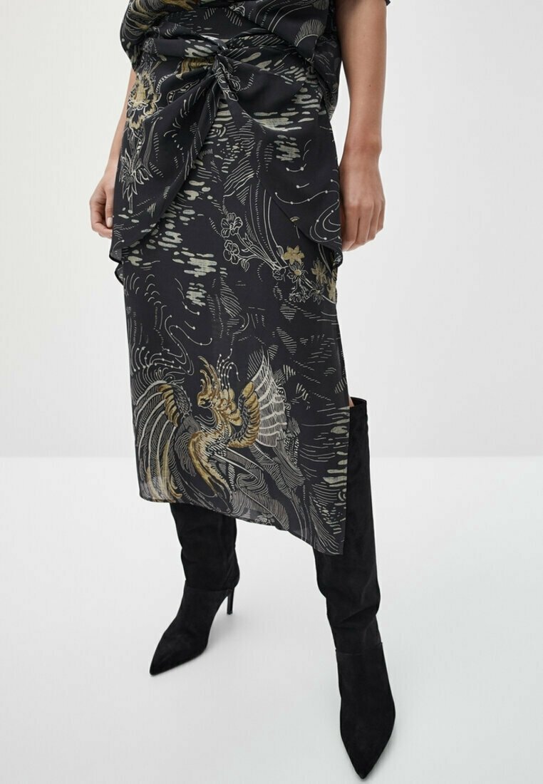 Woman wearing a black and gold patterned wrap skirt with an asymmetrical hem and black suede pointed-toe knee-high boots.