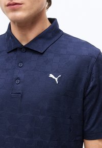 Homme portant un polo bleu marine texturé avec trois boutons et un logo Puma blanc sur le côté gauche de la poitrine.