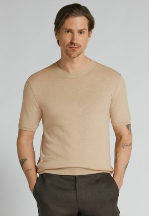 Hombre con cabello de longitud media que lleva un suéter beige de manga corta y pantalones oscuros, de pie con las manos en los bolsillos frente a un fondo liso.