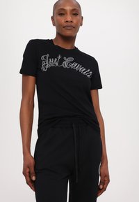 Juodas marškinėlis su blizgančiu sidabriniu "Just Cavalli" logotipu sklandžiu šriftu. Medžiaga turi glotnų tekstūrą, yra prigludusi, su trumpomis rankovėmis.