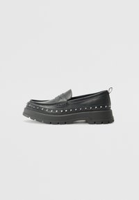 Svarta läderslip-on loafers med kraftiga gummisulor, prydda med pyramidnitar längs kanten och en dekorativ sömsdetalj.