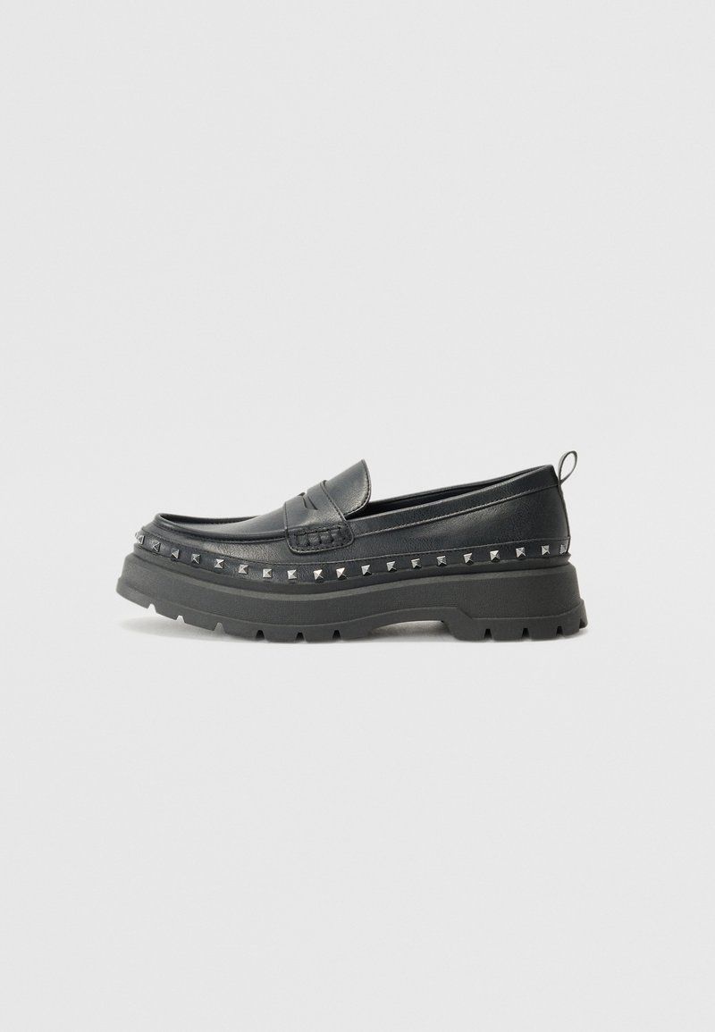 Svarta läderslip-on loafers med kraftiga gummisulor, prydda med pyramidnitar längs kanten och en dekorativ sömsdetalj.