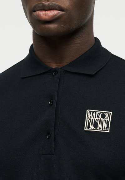 Gros plan sur une personne portant un polo noir à col avec trois boutons et un logo blanc "Maison Kitsuné" sur la poitrine.