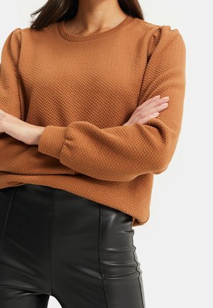 Kobieta ubrana w brązową, fakturowaną bluzkę z długimi, bufkami na rękawach oraz czarne, dopasowane skórzane spodnie, ze skrzyżowanymi ramionami na piersi.