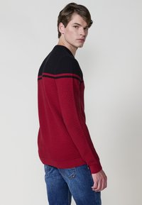 Maglione testurizzato rosso e nero con colletto e polsini a coste, caratterizzato da strisce orizzontali nere sulle spalle. Indossato con jeans blu.