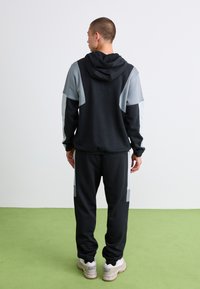 Male wearing black and gray color-block hoodie and matching joggers, standing on green floor facing away from camera.