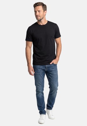 Man draagt een zwart T-shirt met korte mouwen, blauwe spijkerbroek en witte sneakers, staat met één hand in de zak en kijkt naar de zijkant.