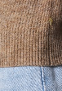 Maglione marrone a coste in lana con dettaglio a forma di albero dorato, abbinato a jeans leggermente blu. Il maglione presenta una stoffa testurizzata in tutto il suo design.