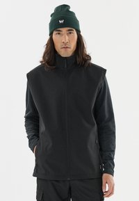 Schwarze ärmellose Jacke mit hohem Kragen, aus glattem Stoff, kombiniert mit einer dunkelgrünen Strickmütze mit Logo.