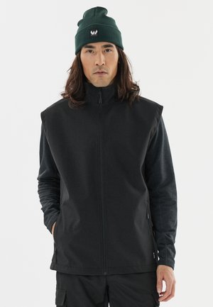 Sort ærmeløs jakke med høj krave, lavet af glat stof, båret sammen med en mørkegrøn strikket beanie med et logo.